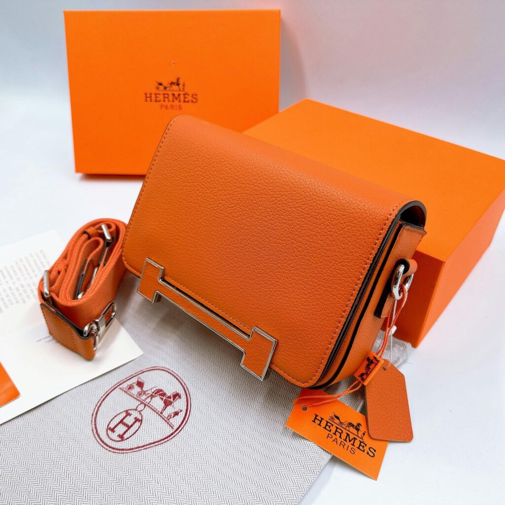 Close-up of an orange Hermes leather handbag with box and tag, showcasing luxury fashion.