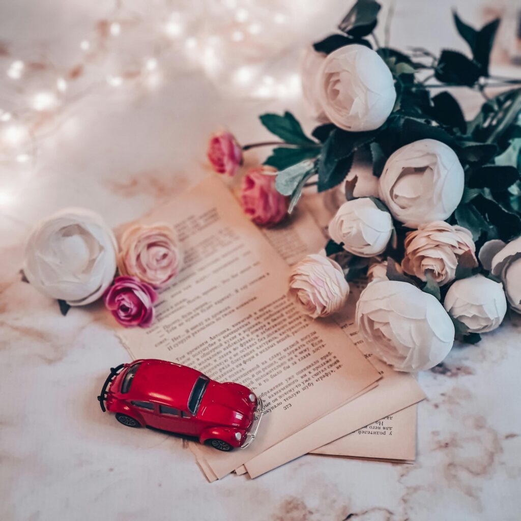 Elegant composition of paper roses, book pages, and a red toy car with fairy lights.